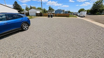 Uma entrada residencial com um carro azul estacionado à esquerda, piso de cascalho e casas ao fundo.