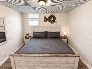 Ein Schlafzimmer mit einem hölzernen Bettgestell, zwei Nachttischen, einer Wanduhr und einem Fenster mit Jalousien.