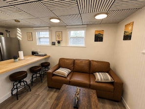 A cozy room with a brown leather sofa, wooden bar stools, a wooden coffee table, and a ceiling with a striped pattern.