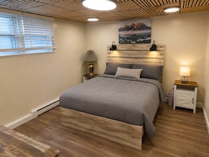 A bedroom with a bed, bedside tables, a lamp, a painting, and a wooden floor.
