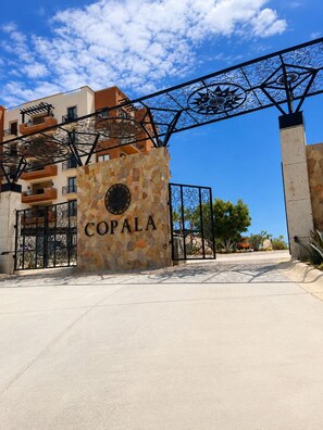 Une entrée fermée par un portail, sur laquelle est inscrit le nom « COPALA » sur un mur de pierre, mène à un bâtiment doté de balcons, avec un ciel dégagé au-dessus.