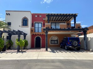 A residential building with a red door, a blue vehicle parked in the driveway, and a pergola structure. - Charming 3-bedroom Modern Mexican house in a Exclusive Copala  Cabo San Lucas (Cabo San Lucas)