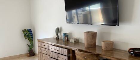 A modern living room with a flat-screen TV mounted on the wall, a wooden desk with drawers, a chair, and a potted plant.