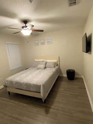 Un dormitorio con una cama, un ventilador de techo, una ventana con persianas y un televisor montado en la pared.