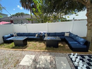 An outdoor seating area with a sectional sofa and two tables, surrounded by a white fence and a tree. - Jax Beach Vacation Stay, 4 bathrooms, 5-bedroom, 2 fire pits, and 2 balconies (Jacksonville Beach)