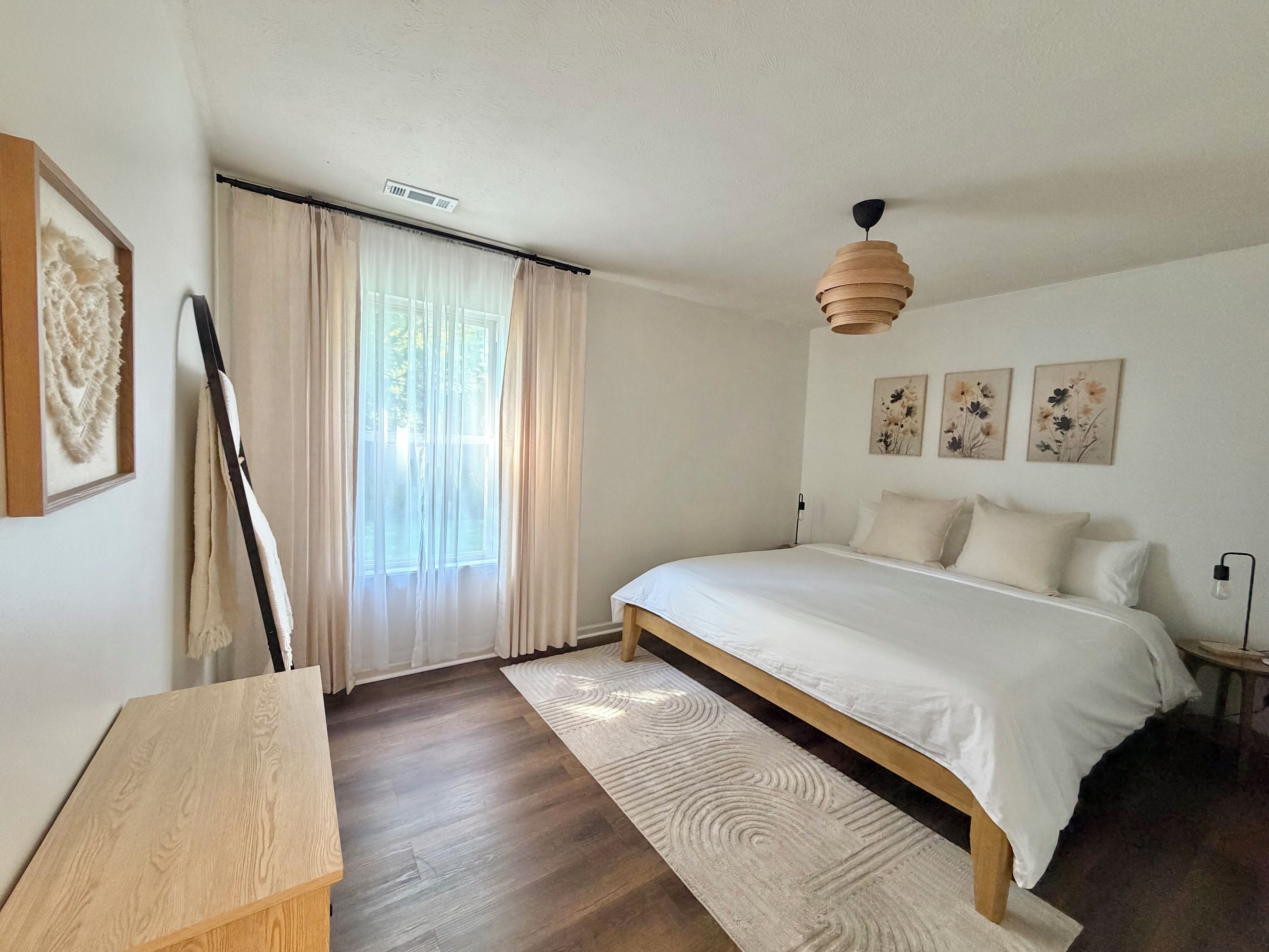 A bedroom with a large bed, a wooden headboard, a bedside table, a floor lamp, a window with sheer curtains, and wall-mounted artwork.