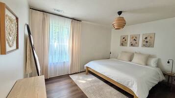 A bedroom with a large bed, a wooden headboard, a bedside table, a floor lamp, a window with sheer curtains, and wall-mounted artwork.