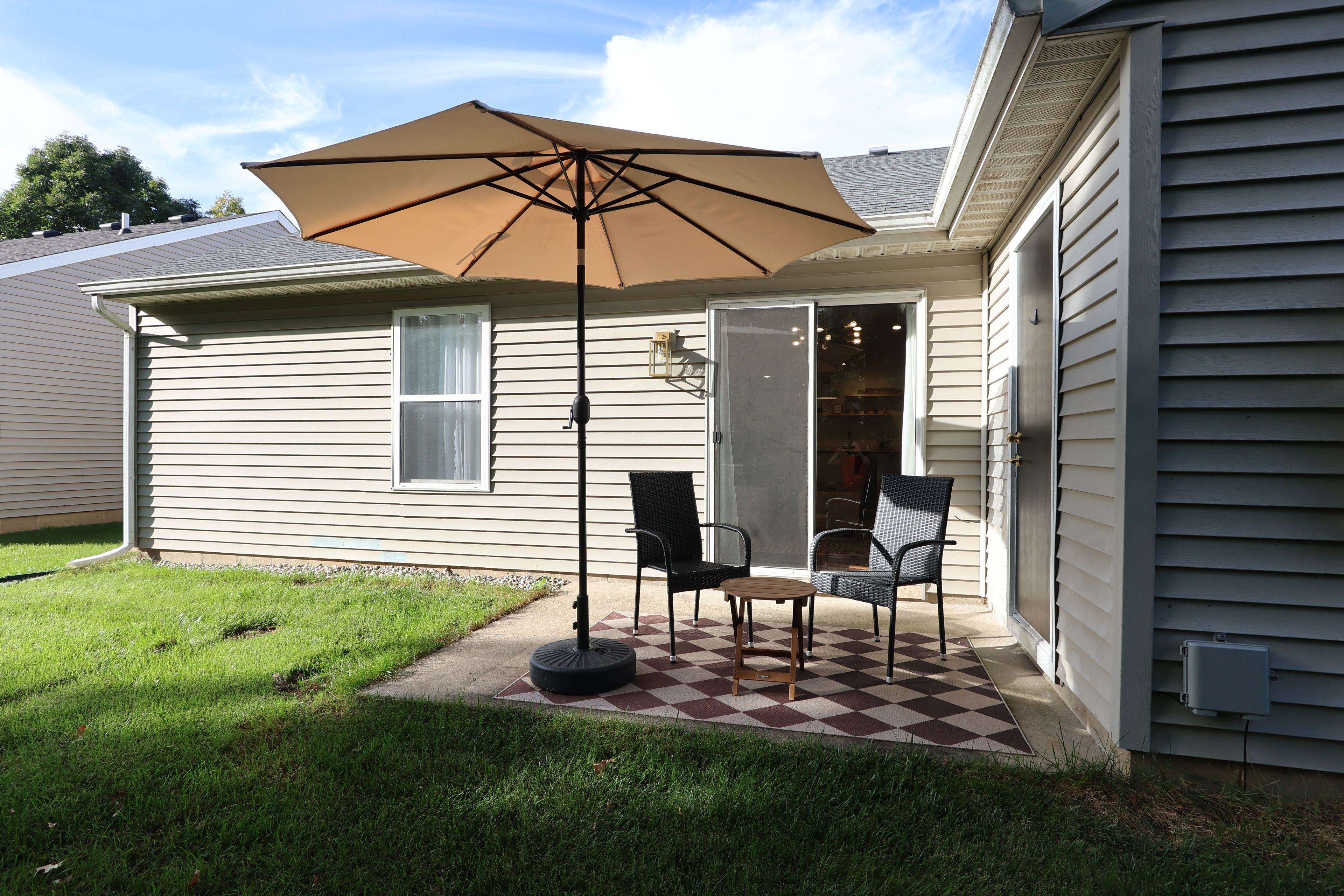 A house with a patio featuring two chairs and a table, a large umbrella, and a checkered floor.