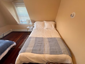 A small, single-bed room with a sloped ceiling, a window with blinds, a bedside table, and a wall-mounted thermostat. - Large 3BR/2BA - 1500sqf - Steps to Harvard (Cambridge)