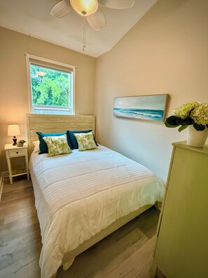 Een slaapkamer met een bed, nachtkastje, raam, schilderij en een bloemenvaas.