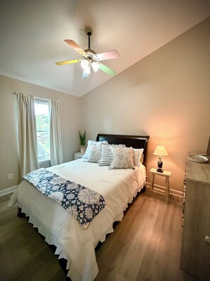 A bedroom with a bed, a nightstand, a lamp, a ceiling fan, and a window with curtains. - Ocean Lakes 3 BR. Golf Cart & Linens. Outdoor Entertainment Area. 3 Min to Beach (Myrtle Beach)