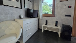 Et værelse med et fjernsyn på en kommode, en stol, et vindue med udsigt og en højttaler.