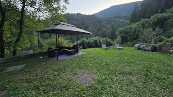 A covered outdoor seating area with a table and chairs, surrounded by lush greenery and a mountainous backdrop.