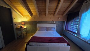 Et soveværelse med træloft, en seng med hvide sengetøj og et rødt tæppe, et natbord med en lampe og et vindue med gardiner.