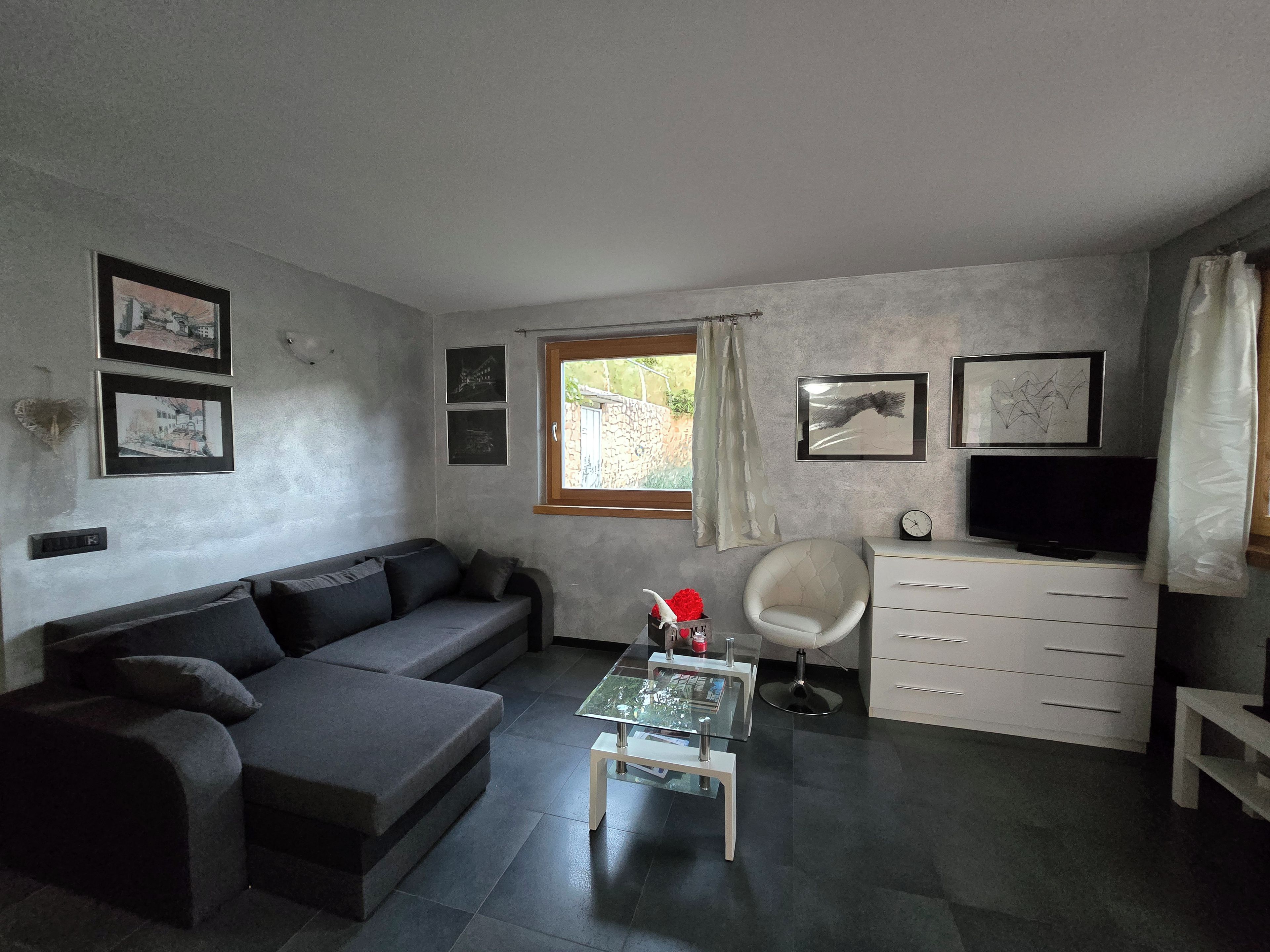 Ein modernes Wohnzimmer mit einem grauen Sofa, einem Glastisch, einem weißen Sessel, einem Fernseher und gerahmten Bildern an der Wand.