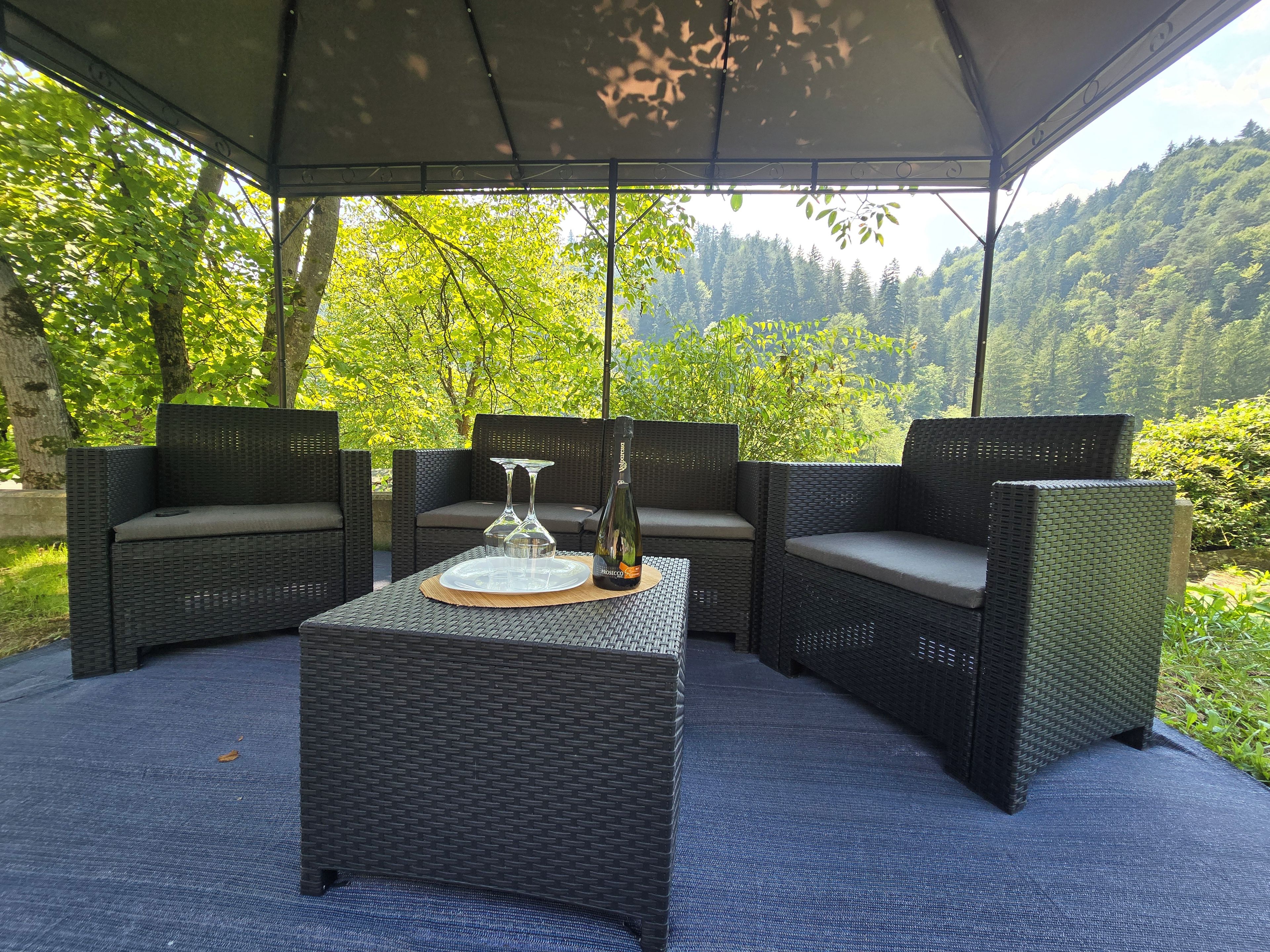 An outdoor seating area with wicker furniture and a table, set against a backdrop of trees and hills.