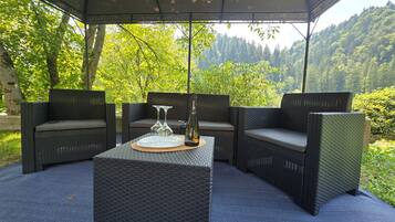 An outdoor seating area with wicker furniture and a table, set against a backdrop of trees and hills.