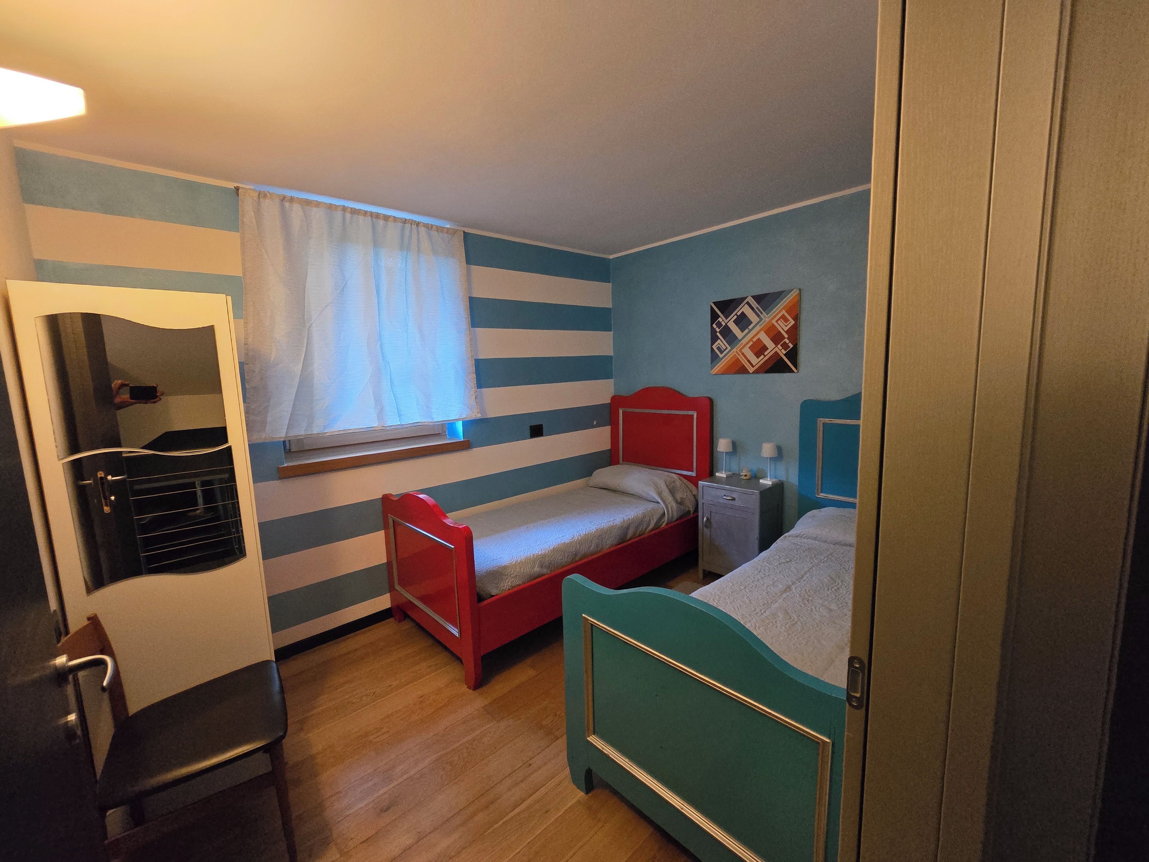 Ein Zimmer mit zwei Betten, einem fensterbelaubten Fenster, einem Spiegel und einer Wand mit geometrischem Muster.