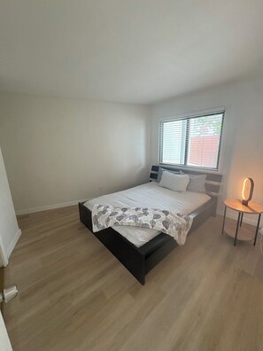 A bedroom with a bed, a bedside table with a lamp, a window with blinds, and a wooden floor. - Luxurious Oceanfront Condo w/ Endless views and Sunsets (San Diego)