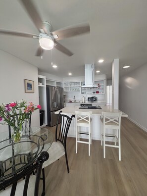 A modern kitchen with a dining area, featuring a glass table, white chairs, and a ceiling fan. - Luxurious Oceanfront Condo w/ Endless views and Sunsets (San Diego)