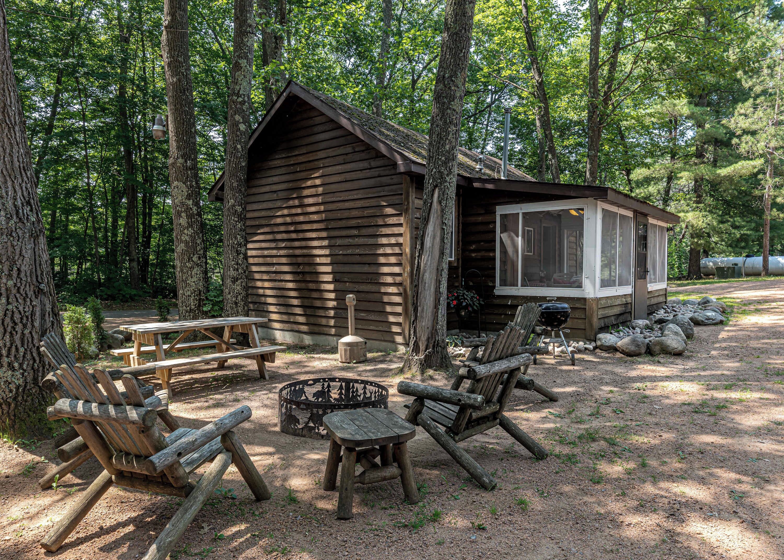 A rustic cabin with a picnic table and two Adirondack chairs in a forest setting.