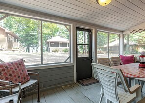 A covered porch with a table and chairs, a door, and large windows overlooking a yard.