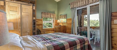 Ein Schlafzimmer mit Bett, Kommode, einem Fenster mit Vorhängen, einem Sessel und Blick ins Freie.