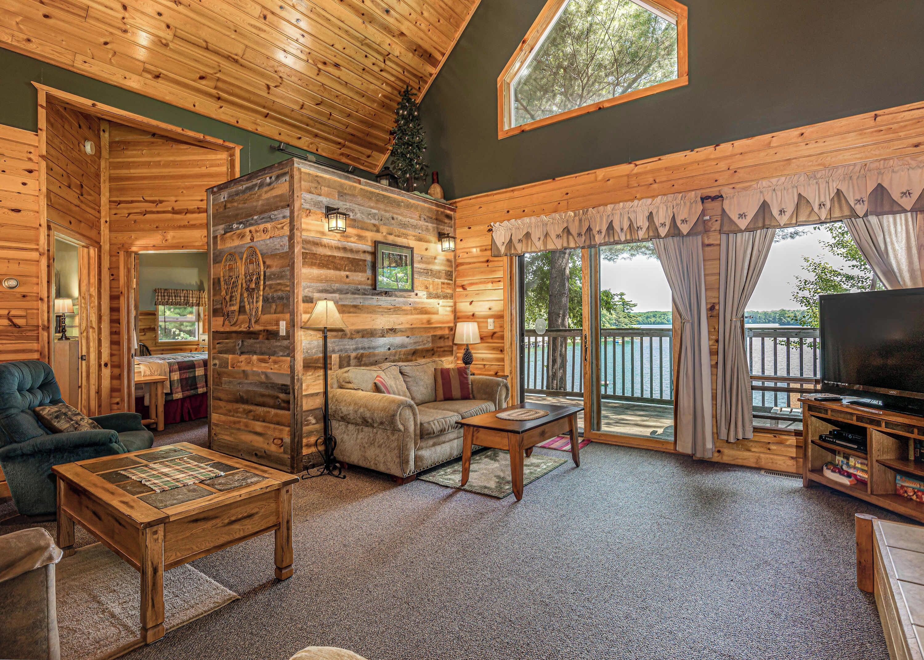 A cozy cabin interior with a wooden floor, a sofa, a coffee table, a TV, and a balcony with a view of the water.
