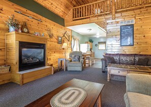 A log cabin interior with a fireplace, wooden furniture, a dining area, and a staircase leading to an upper level. - Maplewood Cabin, Resort, Lake, Beach, Kayaks & More (Lac du Flambeau)