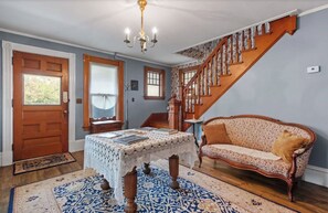 Une pièce avec un escalier en bois, un canapé à motifs, une table en dentelle et un lustre.