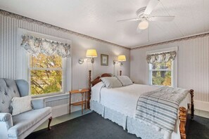 Un dormitorio con cama, una silla, dos lámparas de pared, ventilador de techo y ventanas con cortinas.