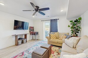 A living room with a sofa, ottoman, television, ceiling fan, and a plant. - Flamingo Hideaway - with Indoor Sauna (Garden City)