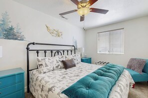 Ein Schlafzimmer mit einem Bett, Deckenventilator, einem Fenster mit Jalousien, einem Nachttisch und einer Kommode.