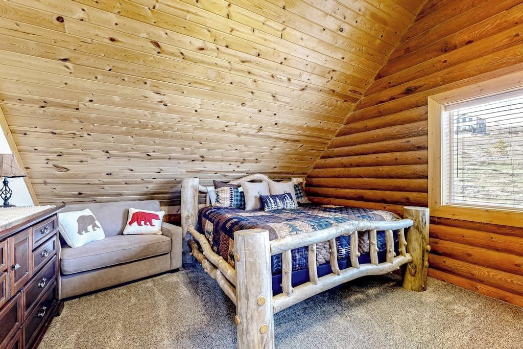 A log cabin bedroom with a wooden ceiling, a bed with a quilt, a sofa with cushions, and a window with a view.