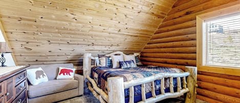 A log cabin bedroom with a wooden ceiling, a bed with a quilt, a sofa with cushions, and a window with a view.