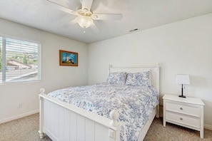 Une chambre avec un lit blanc, une table de chevet, un ventilateur de plafond, une fenêtre avec des stores et un tableau encadré au mur.