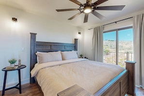 Un dormitorio con cama, ventilador de techo, mesita de noche, una pequeña mesa y ventana con vistas.
