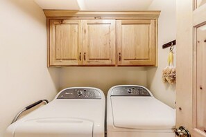 A laundry room with two front-loading washing machines and wooden cabinets above. - Joining Families at Bear Lake (Garden City)