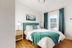 A bedroom with a bed, a nightstand, a lamp, a painting, and a window with curtains. - Secluded Sunrise - Lake Views, Sleeps 23, Dogs OK (Garden City)