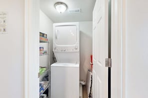 A laundry room with a stacked washer and dryer, a shelf with laundry supplies, and a door with a red handle. - Bear Lake Blue - Lake Views, Heated Pool, Hot Tub (Garden City)