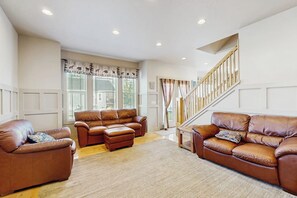 A spacious living room with leather sofas, a wooden staircase, and large windows with curtains. - Bear Shore Retreat - Lake Views, Pool & Game Room (Garden City)