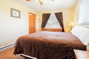A bedroom with a large bed, a wooden nightstand, a window with curtains, and a door. - Meadow Mountain Home (Garden City)