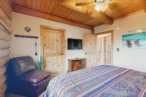 A log cabin room with a bed, a leather chair, wooden doors, a mounted television, and a painting of chairs by the sea. - Elk Ridge Lodge - Log Cabin with Lake Views (Garden City)