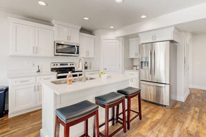 A modern kitchen with white cabinetry, stainless steel appliances, and a central island with bar stools. - Bear Lake Memories - Pickleball & Mountain Views (Garden City)