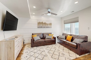 A modern living room with a brown leather sofa, a flat-screen TV mounted on the wall, a ceiling fan, and a large window with blinds. - Bear Lake Memories - Pickleball & Mountain Views (Garden City)