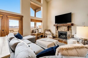 A spacious living room with a stone fireplace, a flat-screen TV, and a large window overlooking a landscape. - Peaceful Bear Lake Home (Garden City)