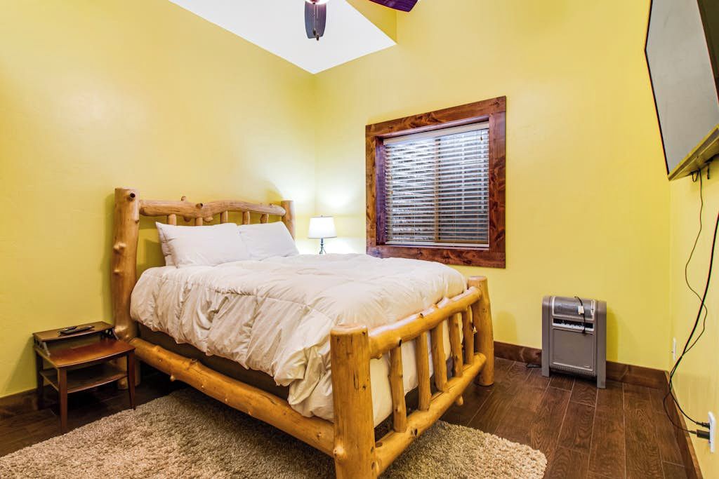 A room with a wooden bed, a small bedside table, a window with blinds, a ceiling light, and a heater.