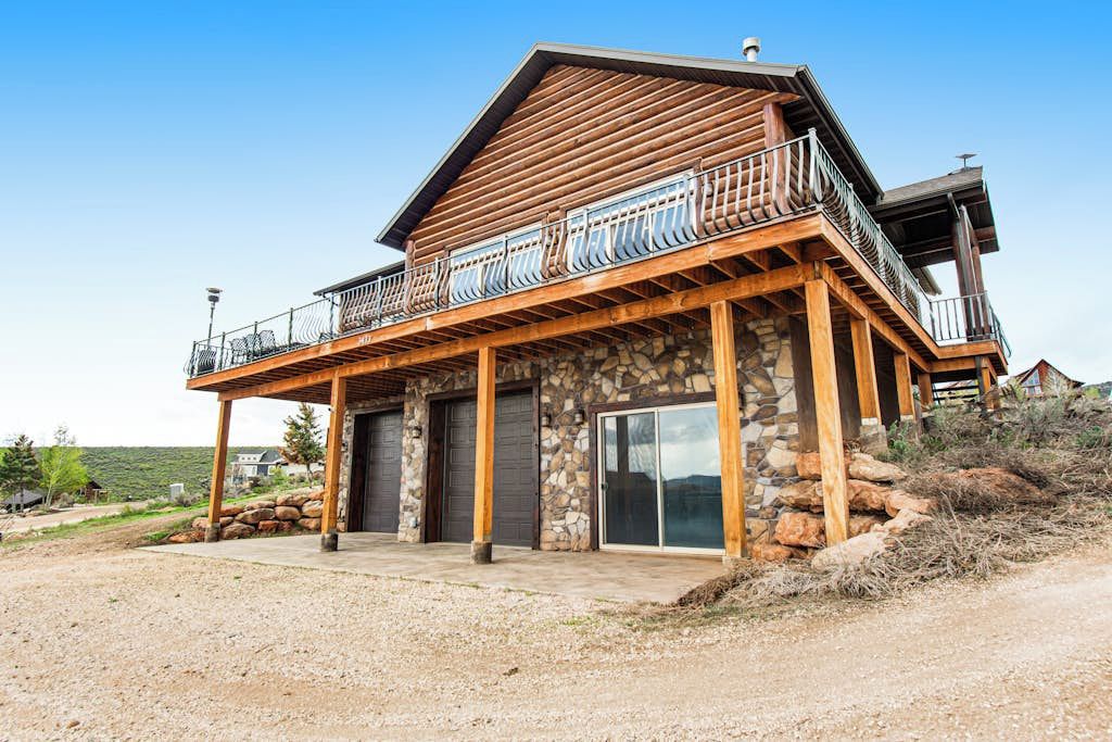 A two-story log cabin with a balcony, stone foundation, and a clear sky.