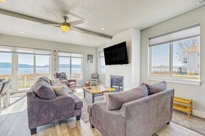 A spacious living room with a fireplace, large windows, and a ceiling fan. - Lakefront Retreat - Hot Tub, Game Room & Fire Pit (Garden City)