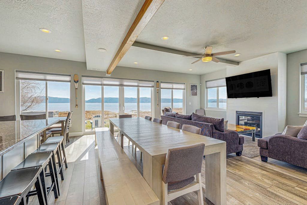 A spacious living area with a long wooden table, multiple seating options, and large windows offering a view of the beach and mountains.